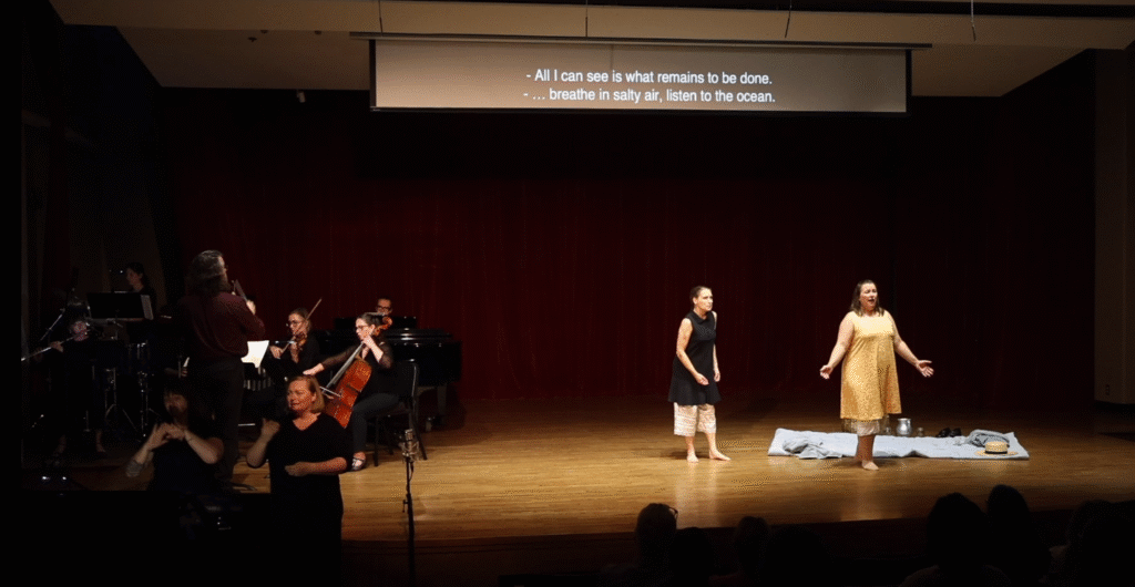 On left, a chamber orchestra, conductor, and two women providing ASL interpretation. On right, MARIE CURIE (Lisa Neher) and IRÈNE CURIE (Lindsey Rae Johnson) sing to one another. 